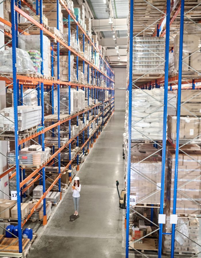 Large warehouse. Woman in white protective helmet with tablet standing in large industrial warehouse in aisle between high racks for goods